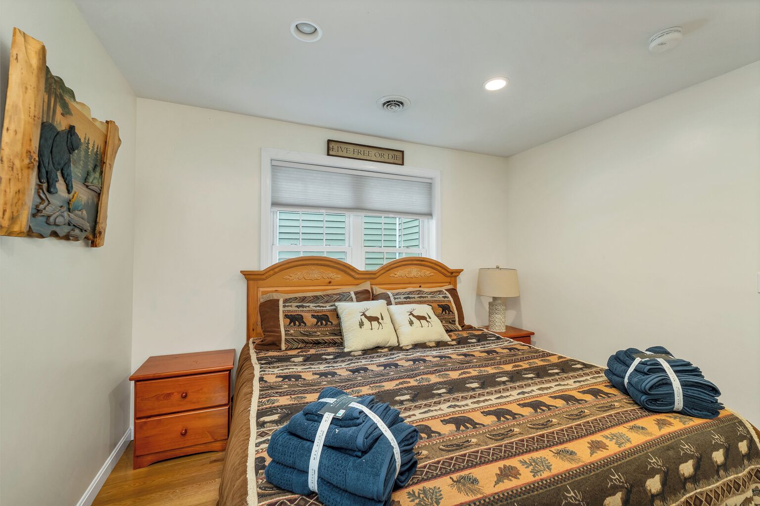 Quarto com cabeceira de madeira, cama com roupa de cama estampada, mesa de cabeceira com abajur e janela com persianas.