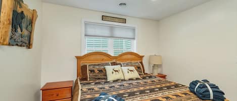 Quarto com cabeceira de madeira, cama com roupa de cama estampada, mesa de cabeceira com abajur e janela com persianas.