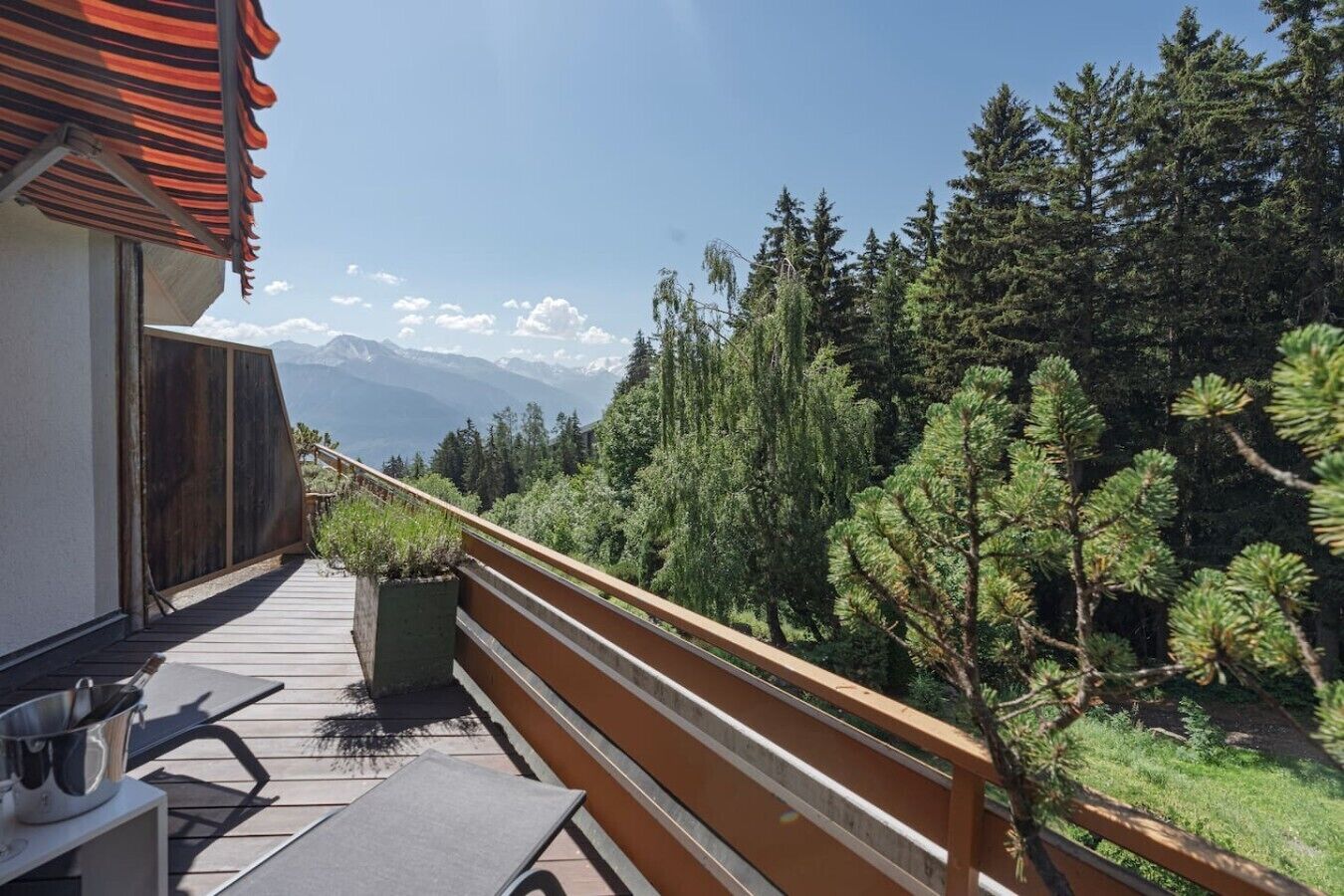 Ein Balkon mit Holzwertkasten, Tisch und Stuhl mit Blick auf eine bewaldete Landschaft und Berge in der Ferne.
