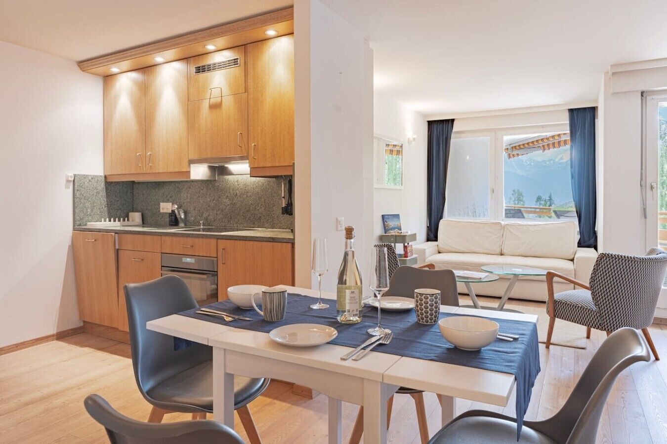 Een moderne keuken met houten kasten, een eettafel gedekt voor twee personen en een zithoek met een bank en uitzicht op de omgeving.
