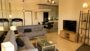 A modern living room with a sofa, coffee table, TV, and a kitchen in the background. - Floisvos Seaview Escape - Fully Equipped Space (Palaio Faliro)