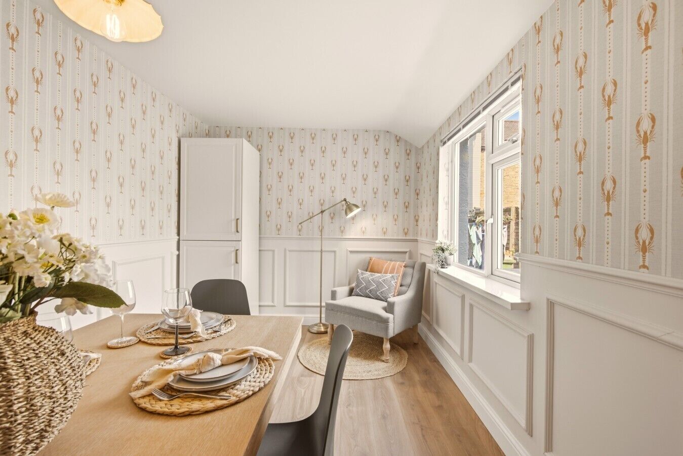 A dining area with a table set for four, a white cabinet, a window with a view, and a patterned wallpaper.