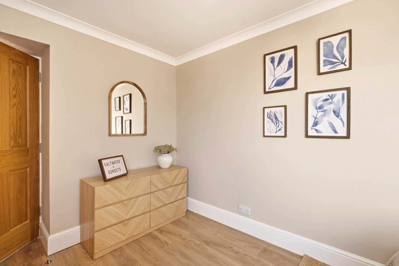 A hallway with a wooden door, a white console table with a plant, and four framed artworks on the wall.