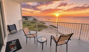 Ein Balkon mit Blick auf den Sonnenuntergang über dem Meer, zwei Stühle, ein kleiner runder Tisch und eine Fußmatte.