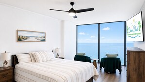 Un dormitorio con cama, mesita de noche, lámpara, ventilador de techo, ventana grande con vista al mar y televisor montado en la pared mostrando 'Dunn Deal Properties & Vacation Rentals'.
