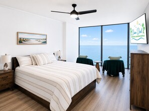 A bedroom with a bed, a nightstand, a lamp, a ceiling fan, a large window with a view of the sea, and a wall-mounted TV displaying 'Dunn Deal Properties & Vacation Rentals'. - Sea Breeze 1004 | Overlooks the Beautiful Gulf (Biloxi)