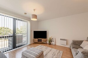 Ein modernes Wohnzimmer mit Flachbildfernseher, einer Couch, einem Couchtisch und einem großen Fenster mit Jalousien.