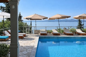 Area piscina con lettini e ombrelloni, vista libera sull'acqua e pavimento in pietra.