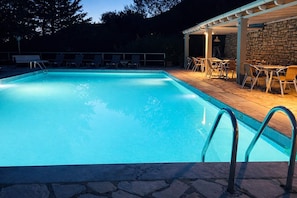 Ein Swimmingpool mit einer Steindeck und Liegestühlen, umgeben von einer Steinmauer und einem überdachten Patio.