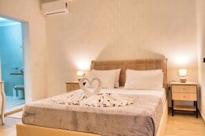 A hotel room with a bed, two bedside tables, a nightstand, a wall-mounted air conditioner, and a bathroom visible through an open door. - Indemela Corfu Lodge  (Karousades)