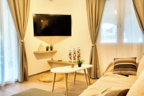 A modern living room with a flat-screen TV mounted on the wall, a sofa with a striped pillow, a small round table with a plant, and a decorative wall shelf. - Indemela Corfu Lodge  (Karousades)