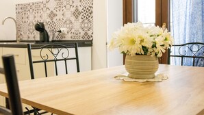 Una mesa de comedor con un jarrón de flores blancas, una cocina con salpicadero estampado y una ventana con cortinas.