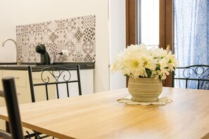 A dining table with a vase of white flowers, a kitchen with patterned backsplash, and a window with curtains. - Mimì's House: Comfort in the Medieval Village of Nettuno (Nettuno)