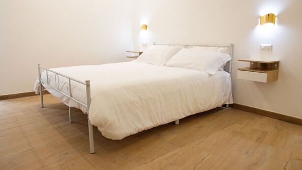 Habitación individual con cama de estructura metálica, ropa de cama blanca, dos mesitas de noche fijadas a la pared y suelo de madera.