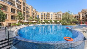 Uma piscina ampla e translúcida em tons de azul, rodeada por um deck ladrilhado com espreguiçadeiras e guarda-sóis.