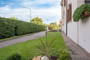 A paved pathway leading to a building with a white facade and brown shutters, flanked by a neatly trimmed hedge and a small garden area with a variety of plants. - SG Family - Il Meriggio (Peschiera del Garda)