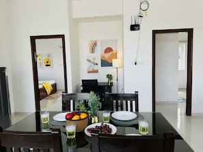 Uma área de refeições com mesa de vidro, cadeiras, uma fruteira, pratos e copos.
