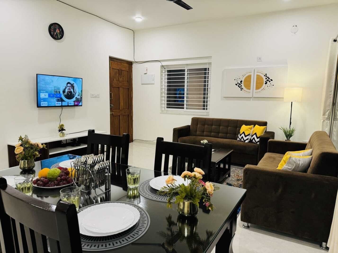 A modern living room with a brown sofa, a dining table set with plates and cutlery, a TV mounted on the wall, and a window with blinds.