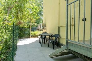 Ein Außenbereich mit Terrasse, einem Tisch, Stühlen, einem grünen Zaun und einem gelben Gebäude.