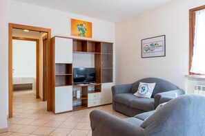 Ein Wohnzimmer mit einer Couch, einem Fernseher und einem Schrank. Eine Tür führt zu einem Schlafzimmer.