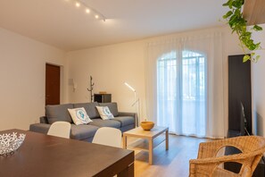 Un salon moderne avec un canapé, une table à manger, un fauteuil en osier et une grande fenêtre avec des rideaux.