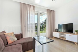 Ein modernes Wohnzimmer mit einer Couch, einem Glastisch, einem Flachbildfernseher auf einem Holz-TV-Möbel und einer Glasschiebetür, die auf einen Balkon führt.