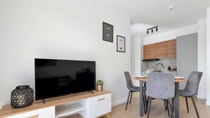 A modern living room with a flat-screen TV on a wooden TV stand, a dining area with a round table and grey upholstered chairs, and a kitchen with wooden cabinets and a black range hood. - Brown Apartment with Garden | Desk | Parking (Jastrzębia Góra)