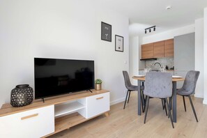A modern living room with a flat-screen TV on a wooden TV stand, a dining area with a round table and grey upholstered chairs, and a kitchen with wooden cabinets and a black range hood. - Brown Apartment with Garden | Desk | Parking (Jastrzębia Góra)