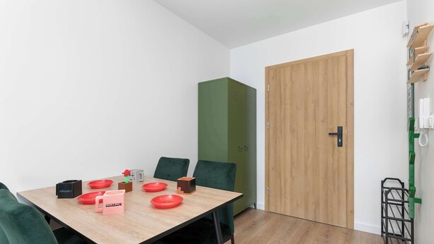 Ein Zimmer mit einem Holztisch, grünen Stühlen, einem grünen Kleiderschrank und einer Holztür.