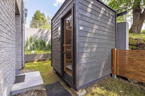 Eine kleine, dunkle Holzhütte mit Glastür, einer Betonbank und einem Holzzaun in einem Garten.