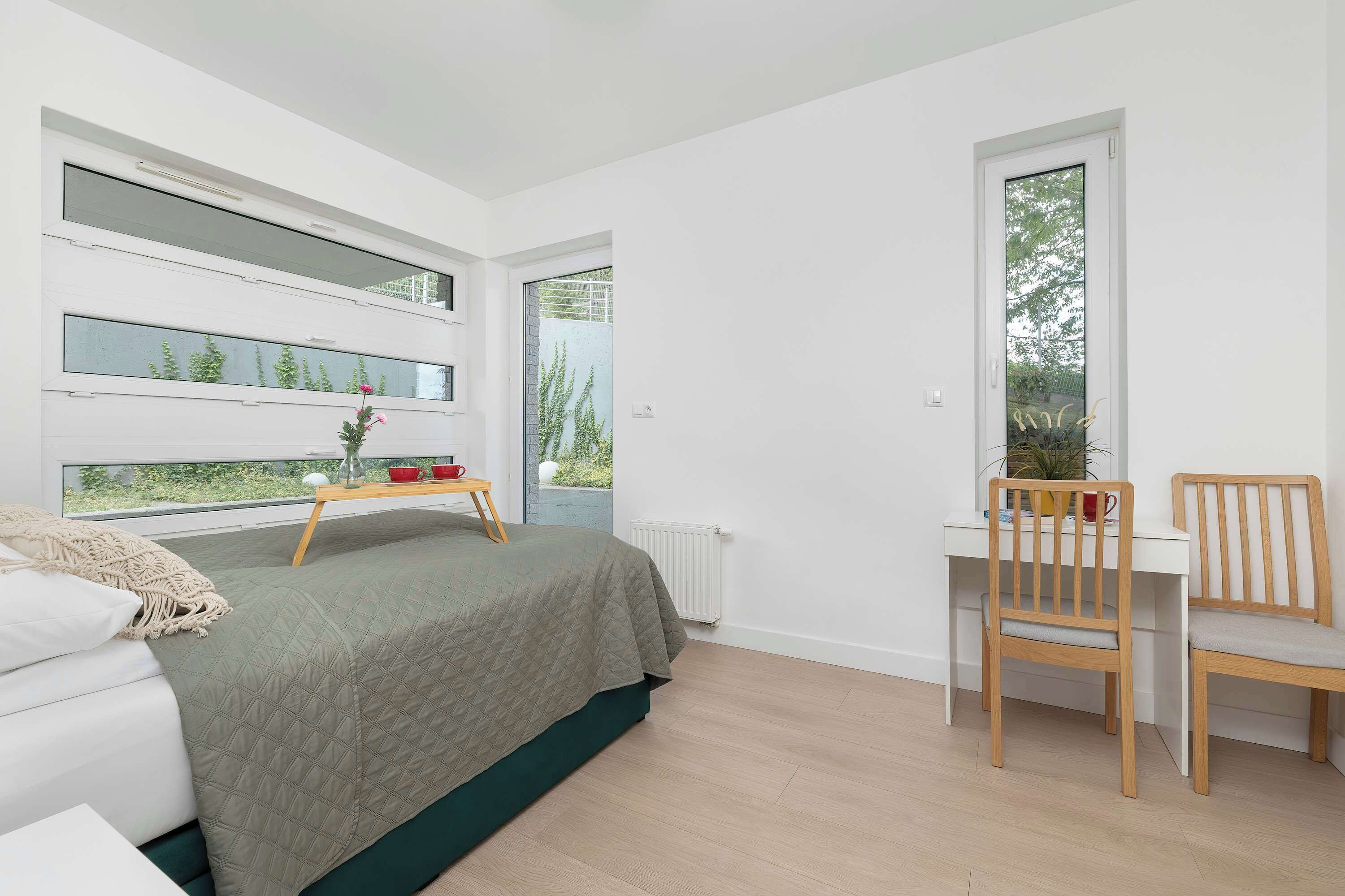 A bedroom with a bed, a desk, and a chair. There are windows with views of greenery.