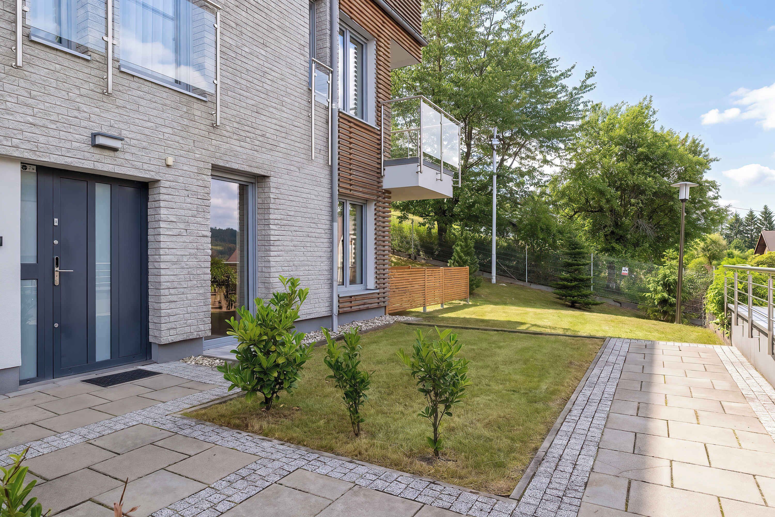 Ein modernes, zweistöckiges Haus mit Backsteinfassade, einer dunklen Haustür und einem kleinen Vorgarten mit jungen Bäumen und einem gepflasterten Weg.