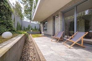 Eine moderne Außenpatio mit zwei Holz-Loungesesseln, einer Betonmauer und einer Glastür zu einem überdachten Bereich.