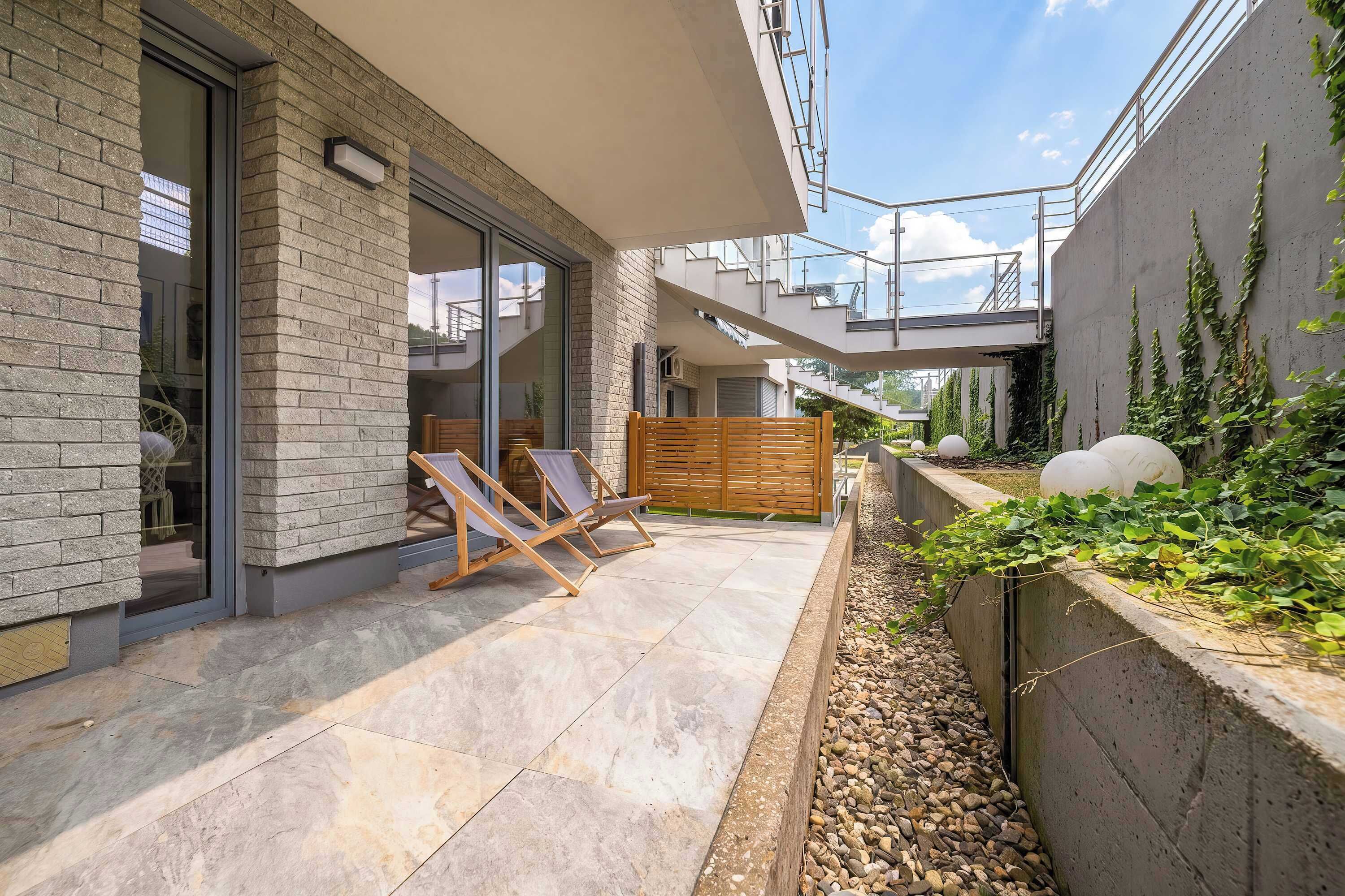 A modern outdoor patio with a stone floor, wooden chairs, and a concrete retaining wall with plants.