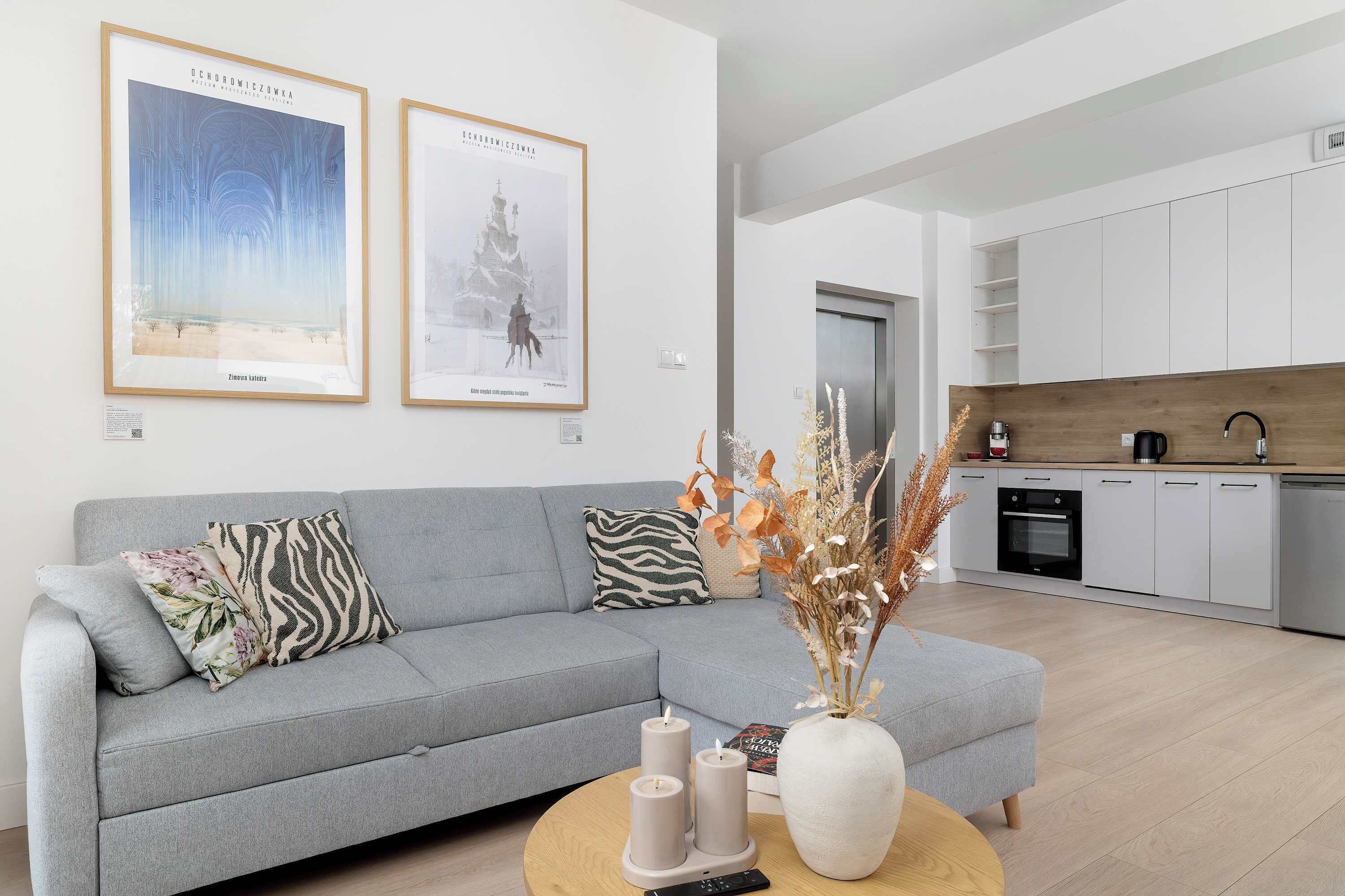 Een moderne woonkamer met een grijze bank, twee ingelijste kunstwerken, een vaas met gedroogde planten en een salontafel.
