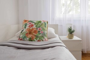 Ein Bett mit einem floralen Kissen, ein Nachttisch mit einer Pflanze und transparente Vorhänge.