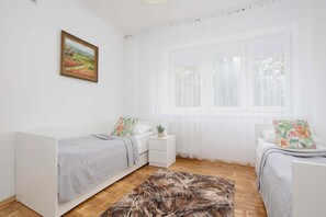 A bedroom with two single beds, a bedside table with a plant, a window with sheer curtains, and a painting on the wall. - Family Apartment for 8 People | Close to the Park (Warszawa)