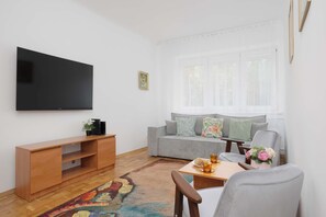 A living room with a flat-screen TV mounted on the wall, a grey sofa with floral cushions, a wooden coffee table, and a patterned rug. - Family Apartment for 8 People | Close to the Park (Warszawa)