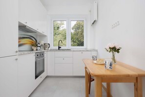 A modern kitchen with white cabinets, a built-in oven, a double sink, and a window with a view of trees. - Family Apartment for 8 People | Close to the Park (Warszawa)