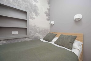 Quarto com cama, duas almofadas, cabeceira, prateleira na parede e luminária fixada na parede.