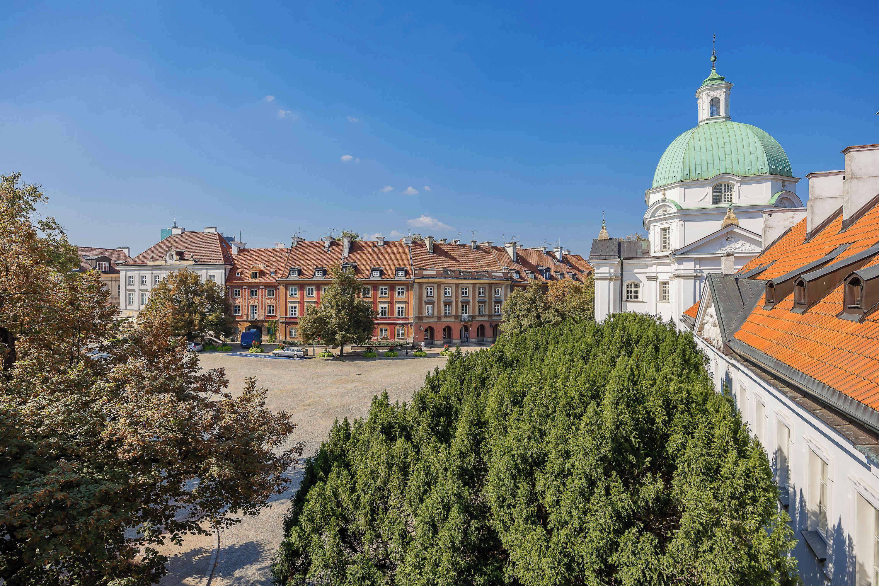 Ein historisches Gebäude mit einer grünen Kuppel und roten Ziegeldächern, umgeben von Bäumen und einem gepflasterten Platz.