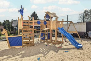 Eine Holzspielkonstruktion mit Rutsche und Kletterelementen, auf sandigem Untergrund, im Hintergrund ein Zaun.