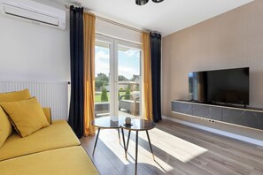 Ein modernes Wohnzimmer mit einer Couch, einem Glastisch, einem Flachbildfernseher und einer Schiebetür zu einem Außenbereich.