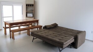 A room with a wooden table, a bench, a sofa, and a built-in shelving unit with a microwave. - Lumina Riviera Ferrugem (Garopaba)
