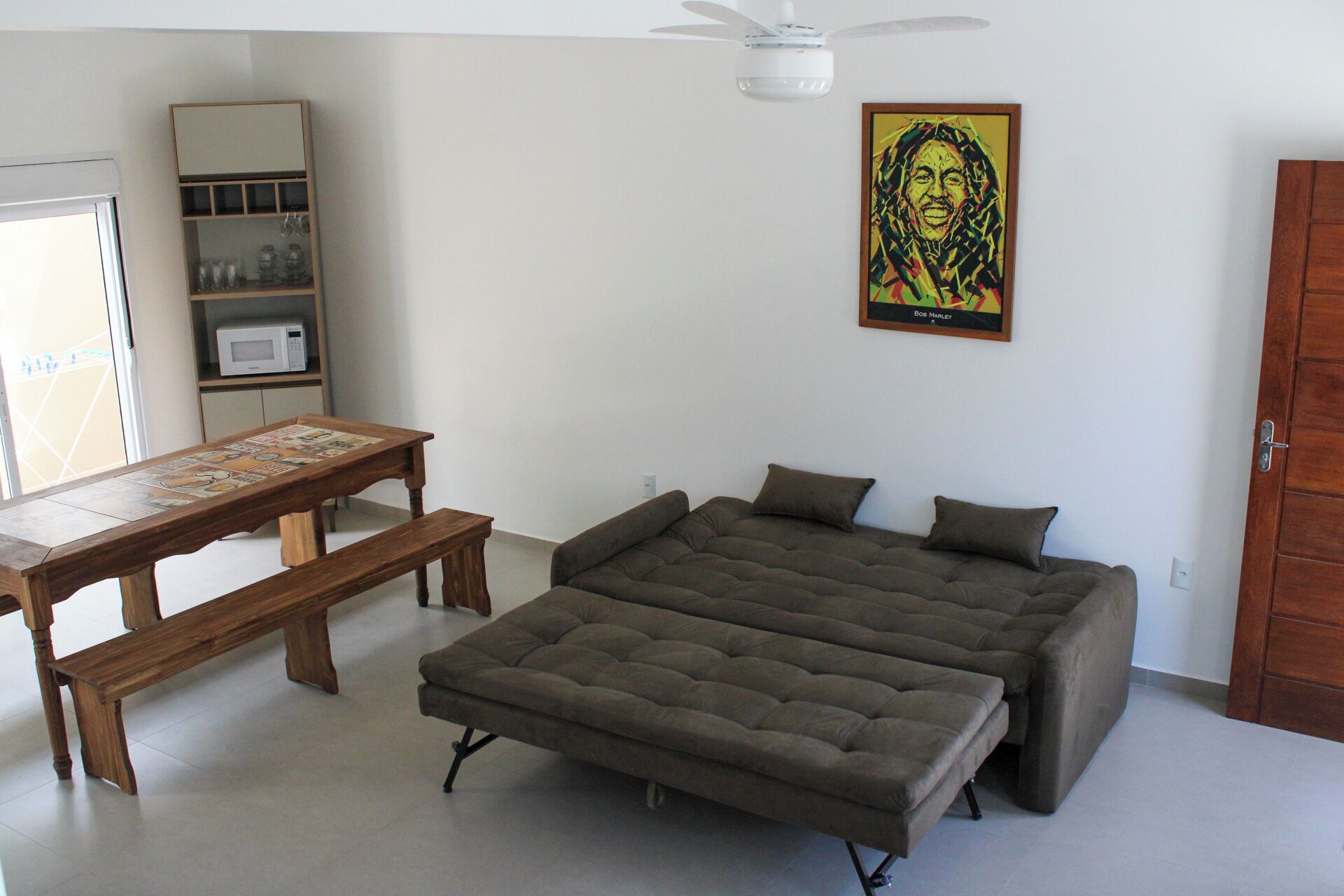 Un dormitorio con cama, mesa, banco, ventilador de techo, microondas y estante.