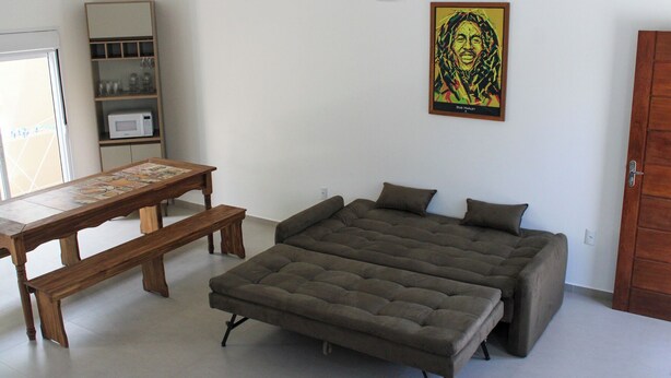 A bedroom with a bed, a table, a bench, a ceiling fan, a microwave, and a shelf. - Lumina Riviera Ferrugem (Garopaba)