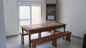A wooden table with a bench in a room with a white door and a shelf with kitchen items. - Lumina Riviera Ferrugem (Garopaba)