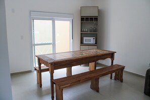 A wooden table with a bench in a room with a white door and a shelf with kitchen items. - Lumina Riviera Ferrugem (Garopaba)