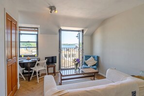 Ein Zimmer mit Esstisch, blauem Sessel, einer Couch und Blick ins Freie durch ein großes Fenster.