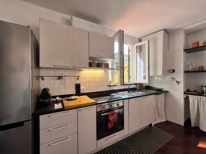 A modern kitchen with white cabinets, a stainless steel refrigerator, a built-in oven, and a window providing natural light. - CASA DEL POZZO (Rapallo)